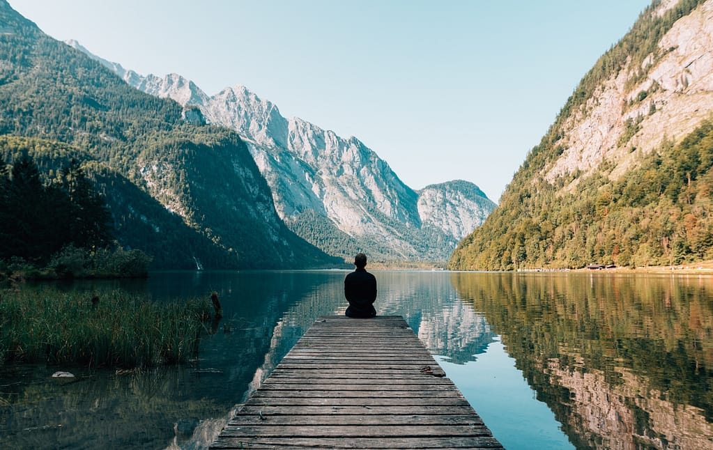 Man sitting at the end of a dock, overlooking a peaceful lake surrounded by mountains