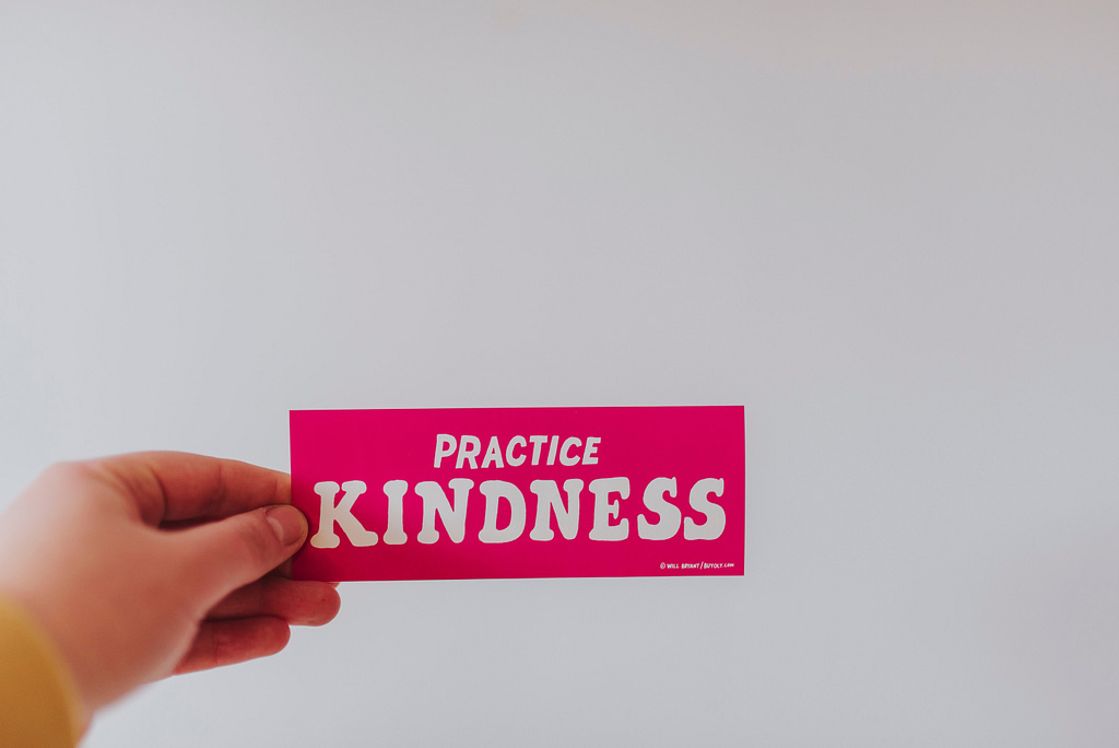A hand appears at the bottom left of the page holding a pink "Practice Kindness" banner
