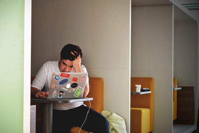 Man sitting in a booth looking at his computer, overwhelmed, with hand on his head. Overcome The Feeling Of Overwhelm